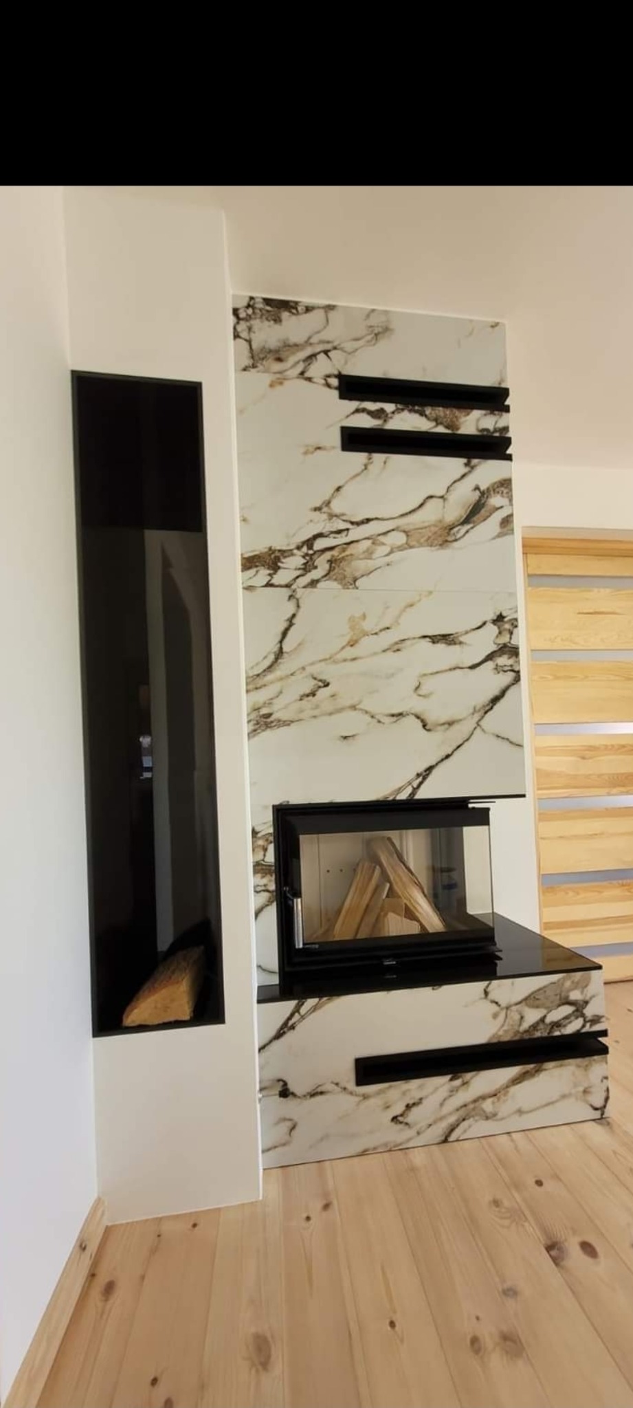 Nowoczesny kominek z obudową z płytek imitujących marmur, z czarnymi wnękami i szybą paleniska, drewno przygotowane do palenia, drewniana podłoga.