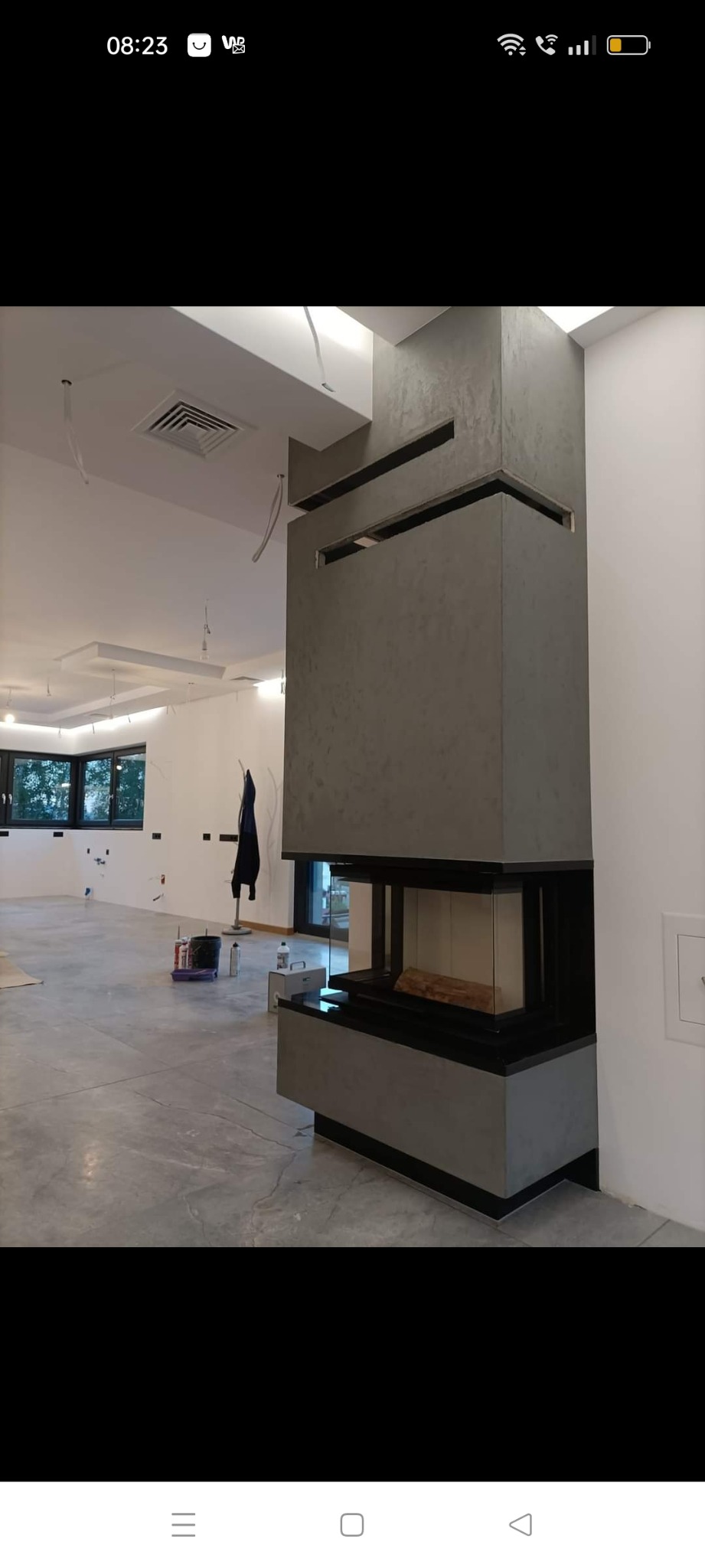 Nowoczesny kominek z obudową z szarego betonu architektonicznego, z widocznym drewnem w palenisku, umieszczony w przestronnym wnętrzu w trakcie wykańczania.