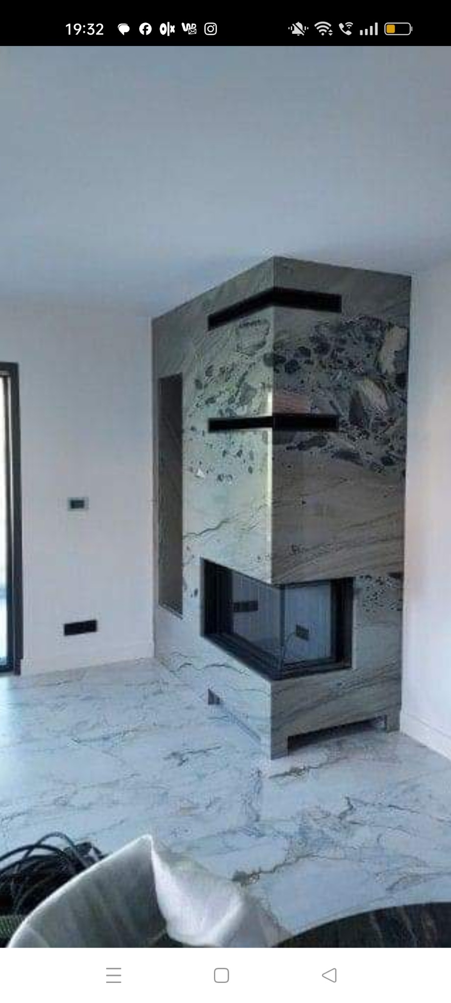 Nowoczesny kominek narożny z obudową z szarego marmuru z czarnymi akcentami, w trakcie montażu w nowym pomieszczeniu z marmurową podłogą.