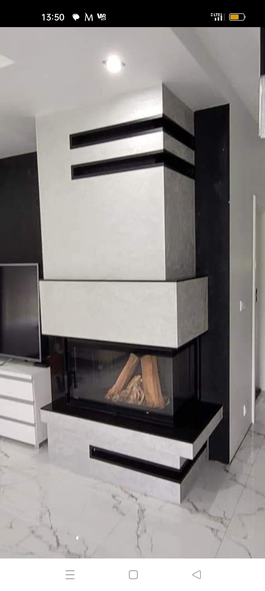 Nowoczesny kominek narożny z obudową w odcieniach szarości i czerni, z widocznym paleniskiem i drewnem wewnątrz, na tle marmurowej podłogi i czarnej ściany.