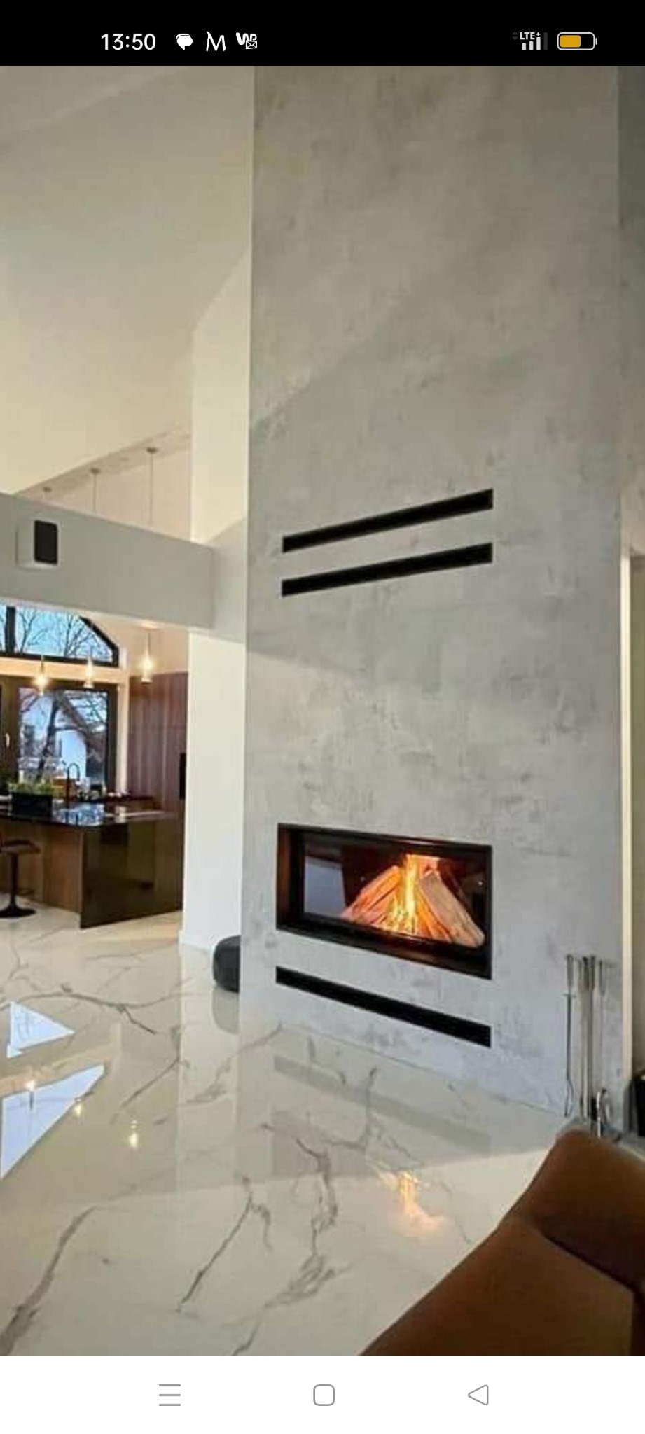 Nowoczesny kominek w zabudowie z szarego betonu architektonicznego, z dwoma poziomymi szczelinami wentylacyjnymi, płomienie widoczne przez szybę, w tle marmurowa podłoga i fragment otwartej kuchni.