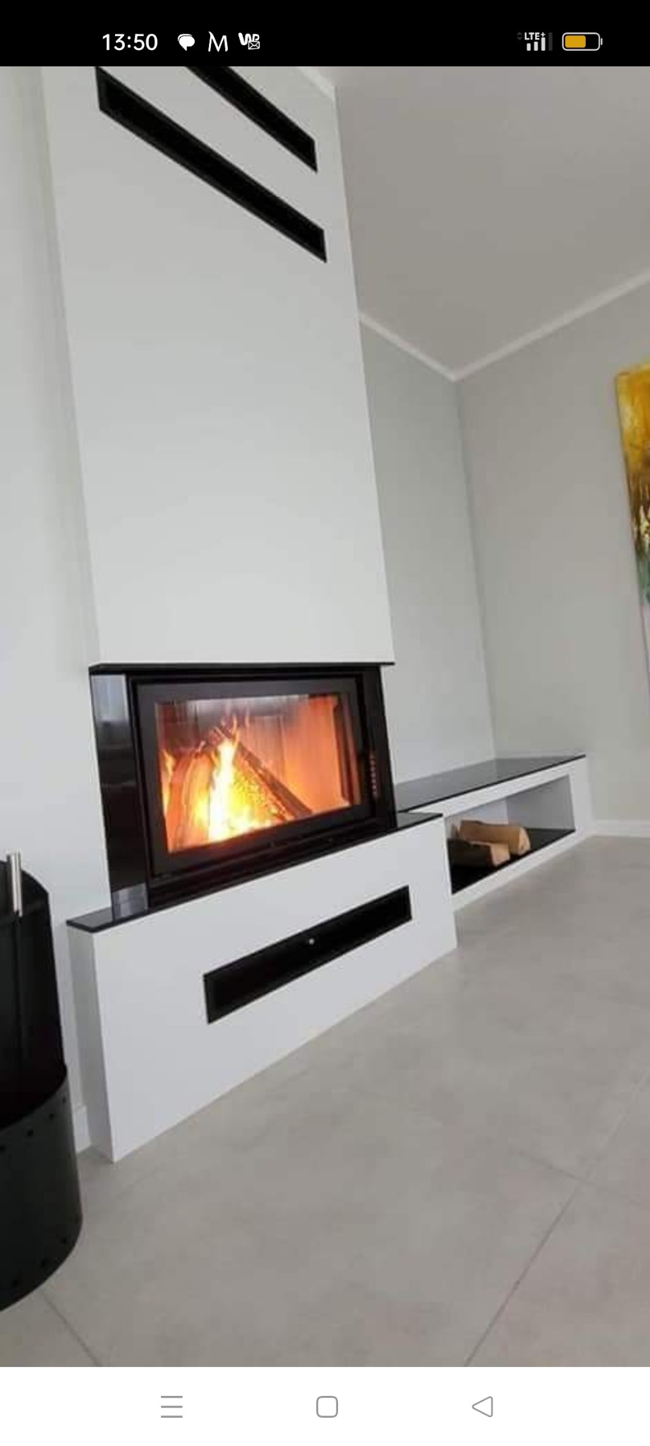 Nowoczesny kominek w zabudowie z białej płyty z czarnymi akcentami i płomieniami wewnątrz paleniska, z półką na drewno obok, na tle szarej ściany i podłogi.