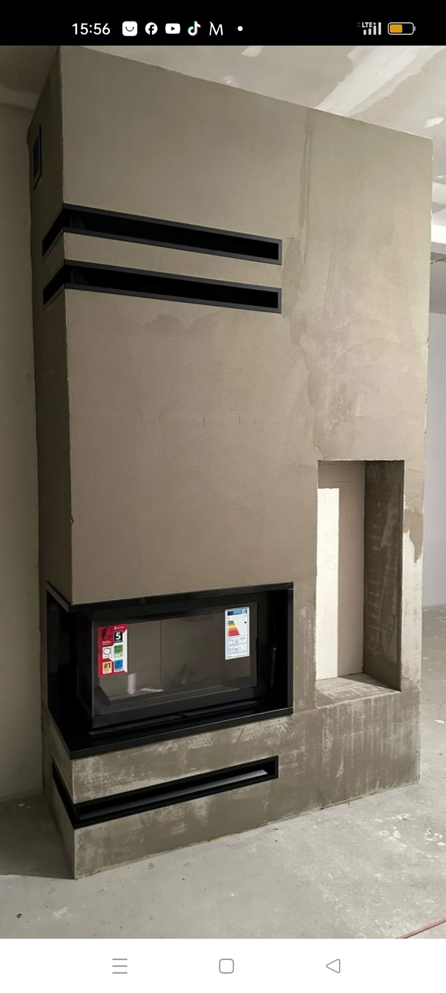 Surowy, wysoki kominek narożny z czarnym wkładem i poziomymi szczelinami wentylacyjnymi w trakcie wykańczania, z widocznymi śladami szpachli i otwartą wnęką obok.