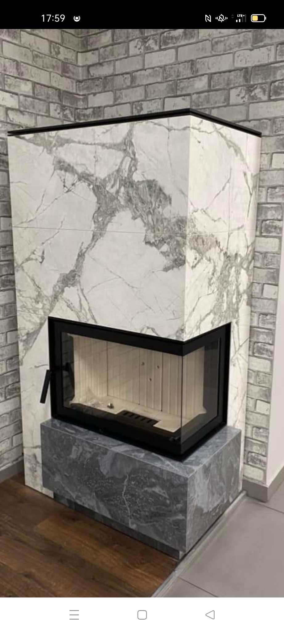 Nowoczesny kominek obudowany płytami z wzorem marmuru, z czarną ramą i drzwiczkami, stojący na podstawie z ciemniejszego marmuru, na tle ściany z szarej cegły.