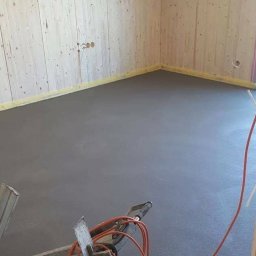 Zbych-bud - Świeżo wylana, gładka, szara posadzka w pomieszczeniu z boazerią, widoczne narzędzia i kable.
