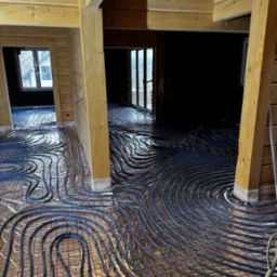 Montaż ogrzewania podłogowego w drewnianym domu, widoczne rury grzewcze rozłożone na folii z siatką.