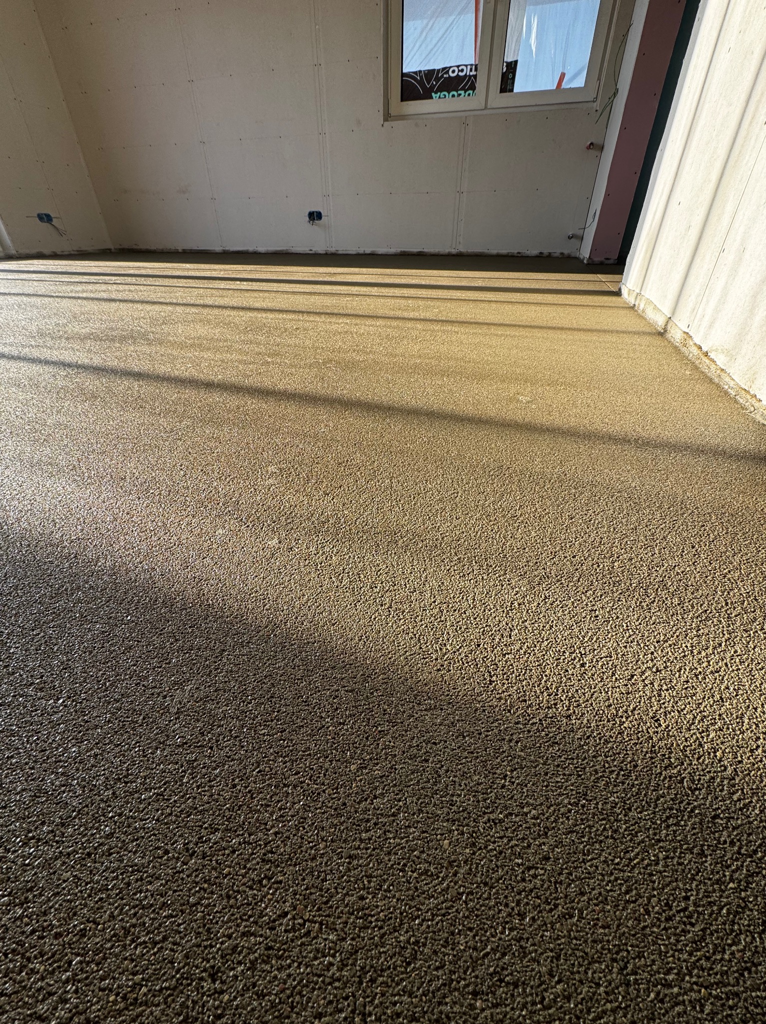 Świeżo wylana, mokra podłoga w surowym stanie, z widocznymi promieniami słońca padającymi przez okno pod skosem.