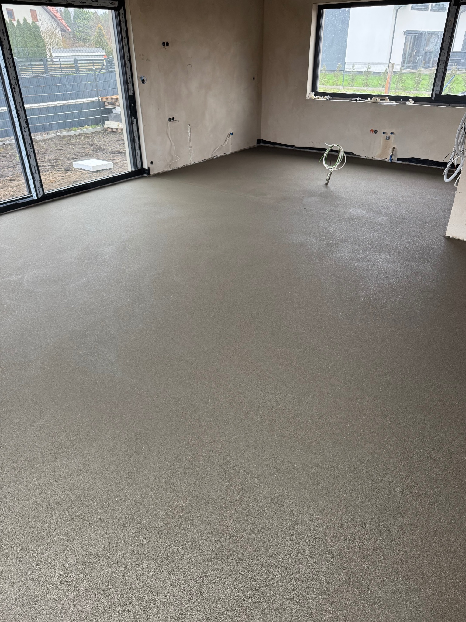 Świeżo wylana, gładka, szara posadzka w surowym wnętrzu z dużymi oknami i wystającymi przewodami elektrycznymi.
