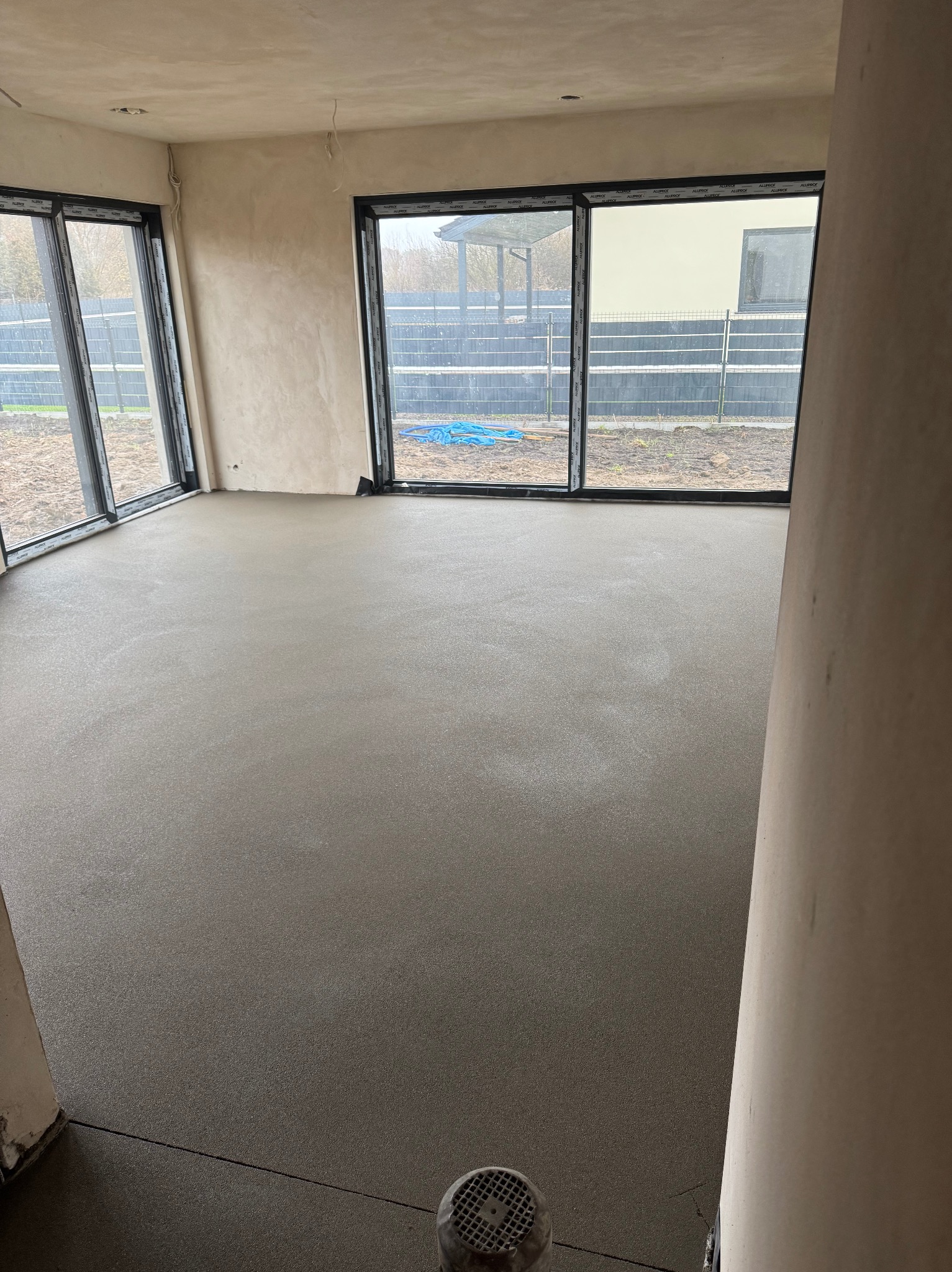 Świeżo wylana, gładka podłoga w surowym stanie, widoczne duże okna z widokiem na ogród, szare ściany w tle i kratka wentylacyjna w rogu.