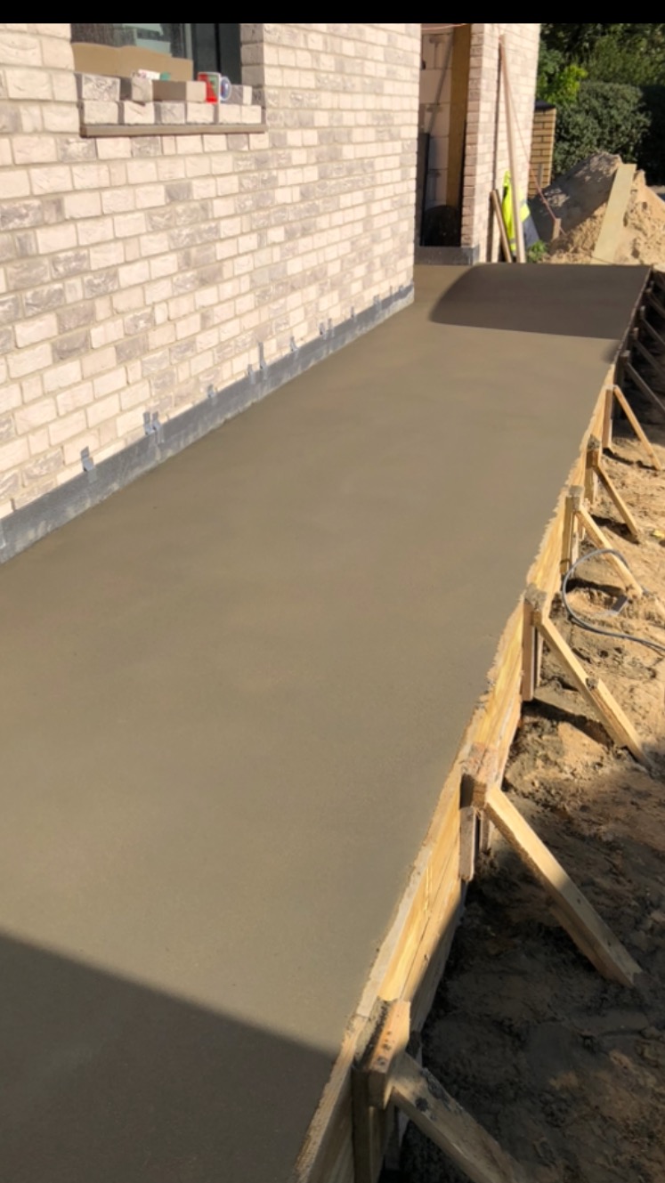 Świeżo wylana, gładka wylewka betonowa z drewnianym szalunkiem, przylegająca do ściany budynku z jasnej cegły.