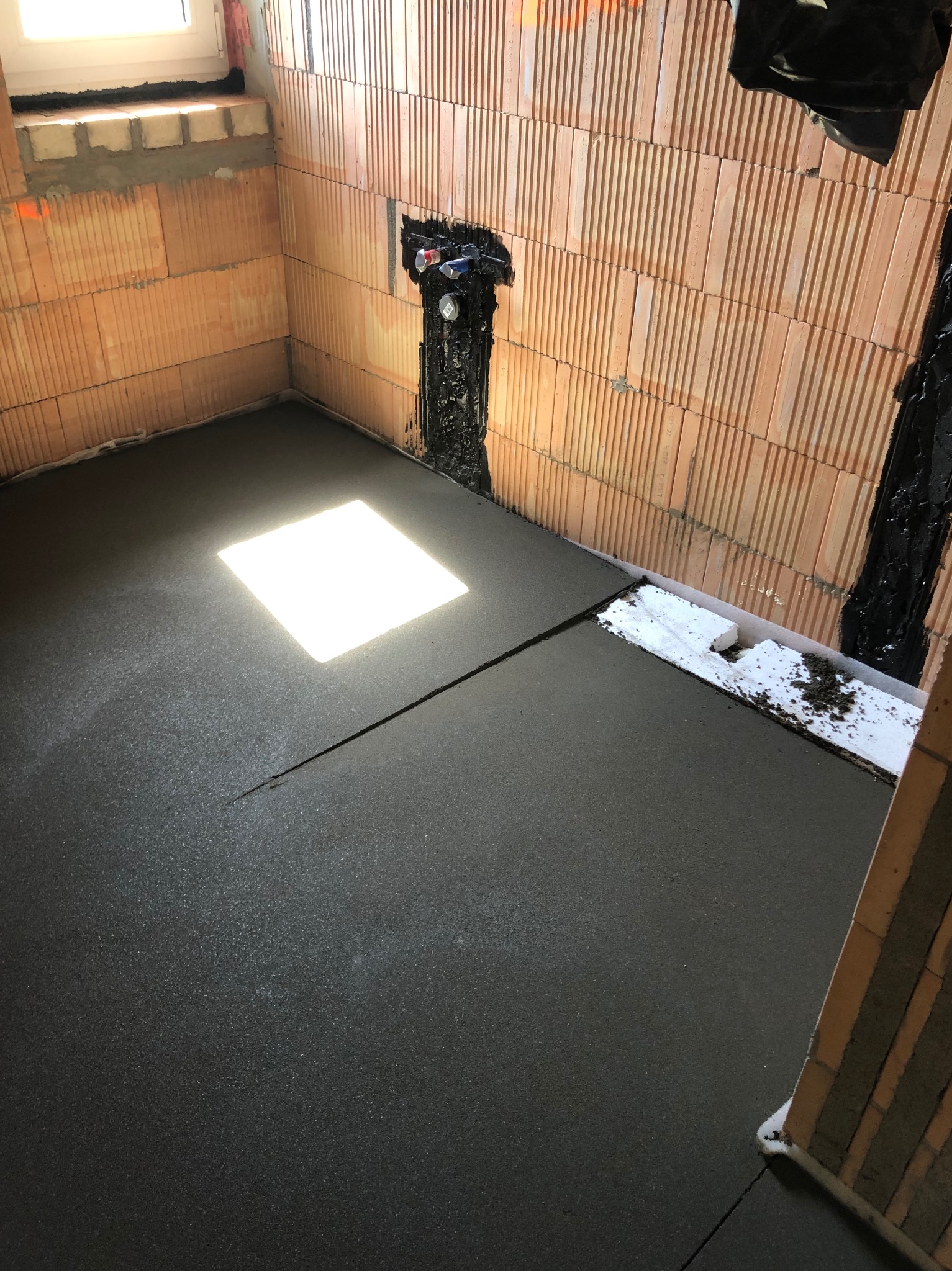 Świeżo wylana, gładka, ciemnoszara wylewka podłogowa w pomieszczeniu z ceglanymi ścianami i widoczną instalacją hydrauliczną.