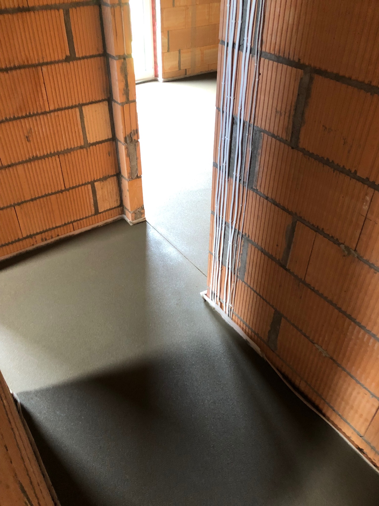 Świeżo wylana, gładka posadzka w surowym wnętrzu z ceglanymi ścianami i pionowymi rurami wzdłuż framugi drzwi.
