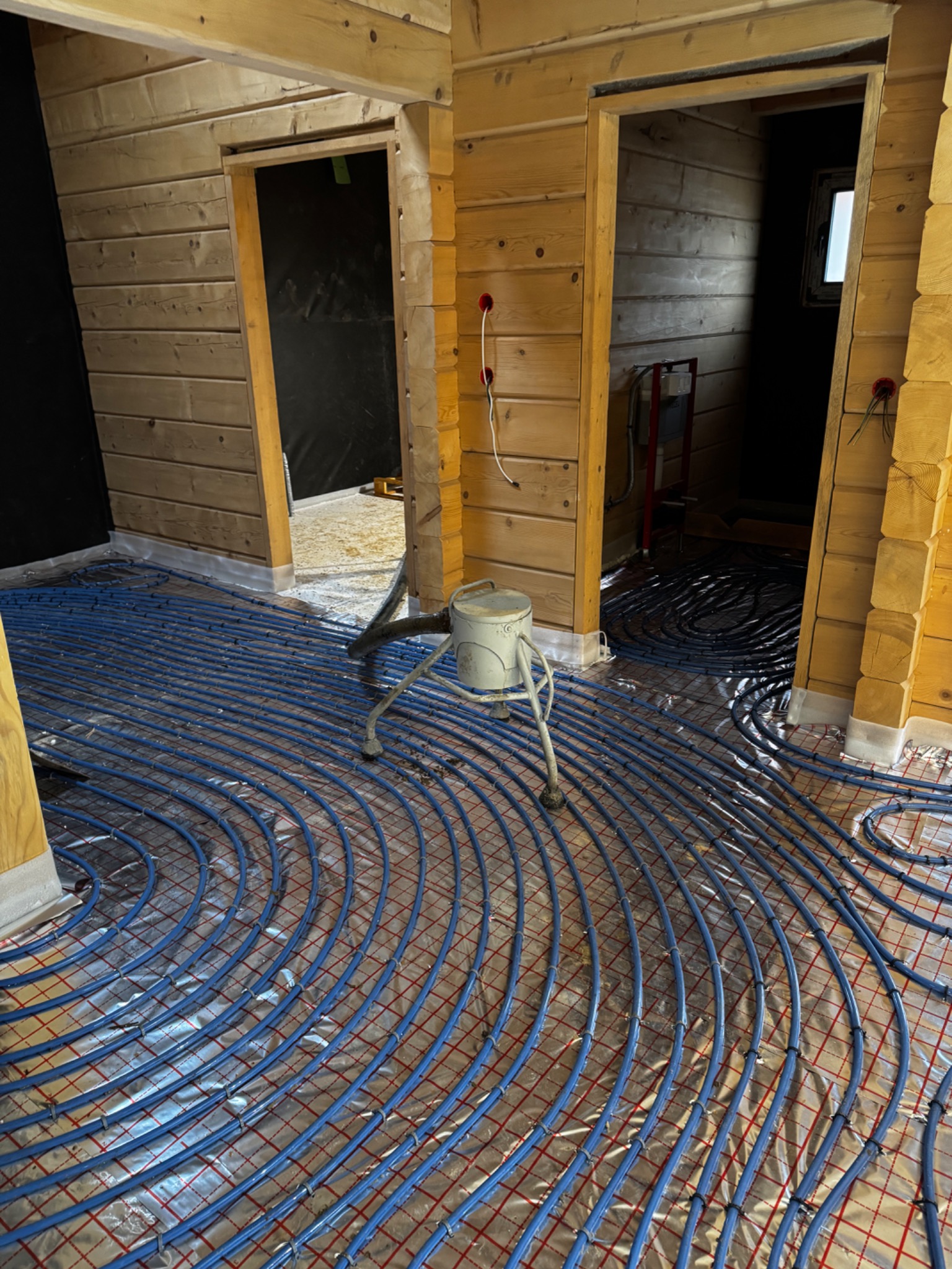 Instalacja ogrzewania podłogowego z niebieskich rurek na folii aluminiowej w drewnianym domu, widoczne dwa wejścia do pomieszczeń.