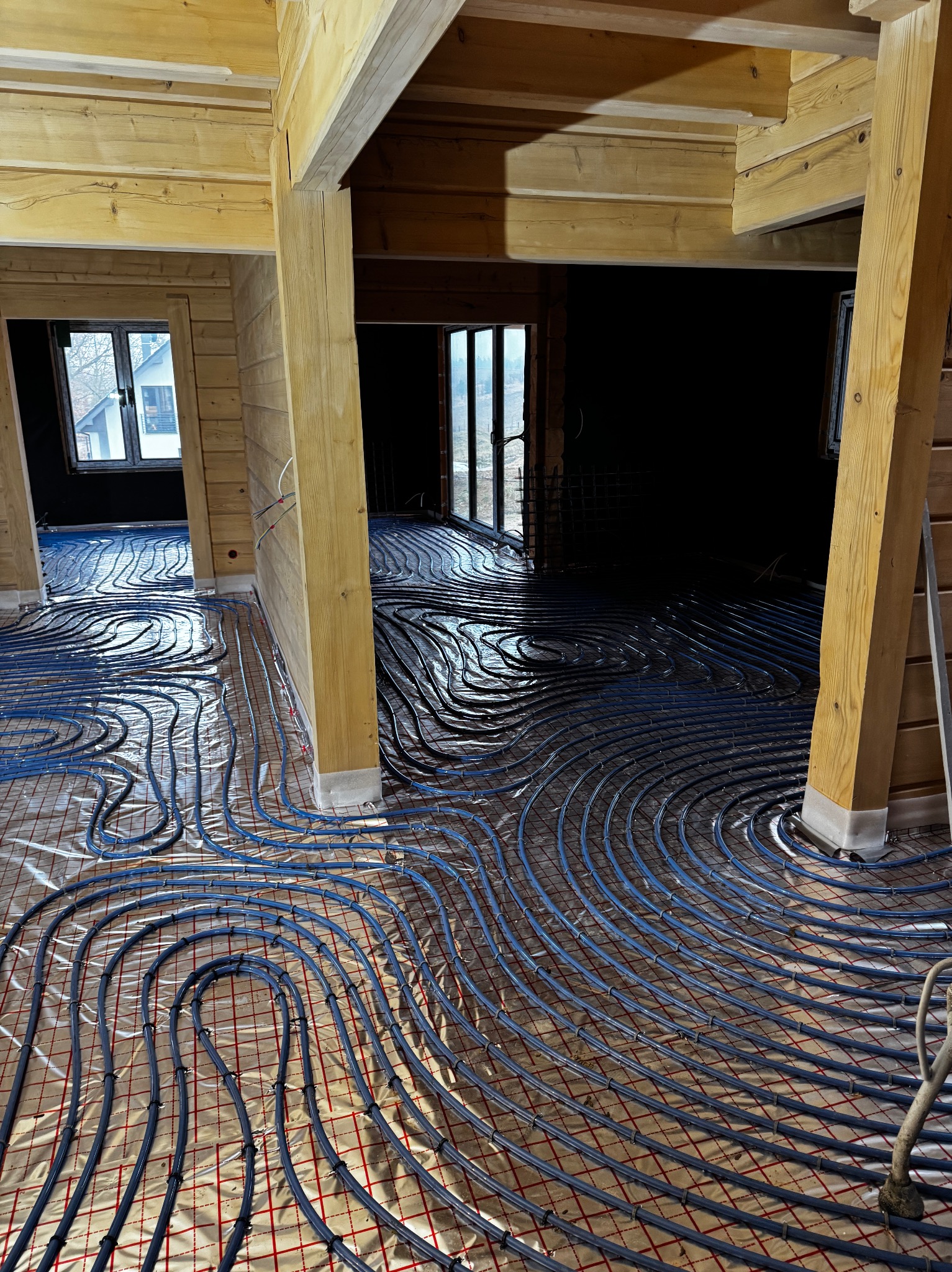 Montaż ogrzewania podłogowego w drewnianym domu, widoczne rury grzewcze rozłożone na folii z siatką.