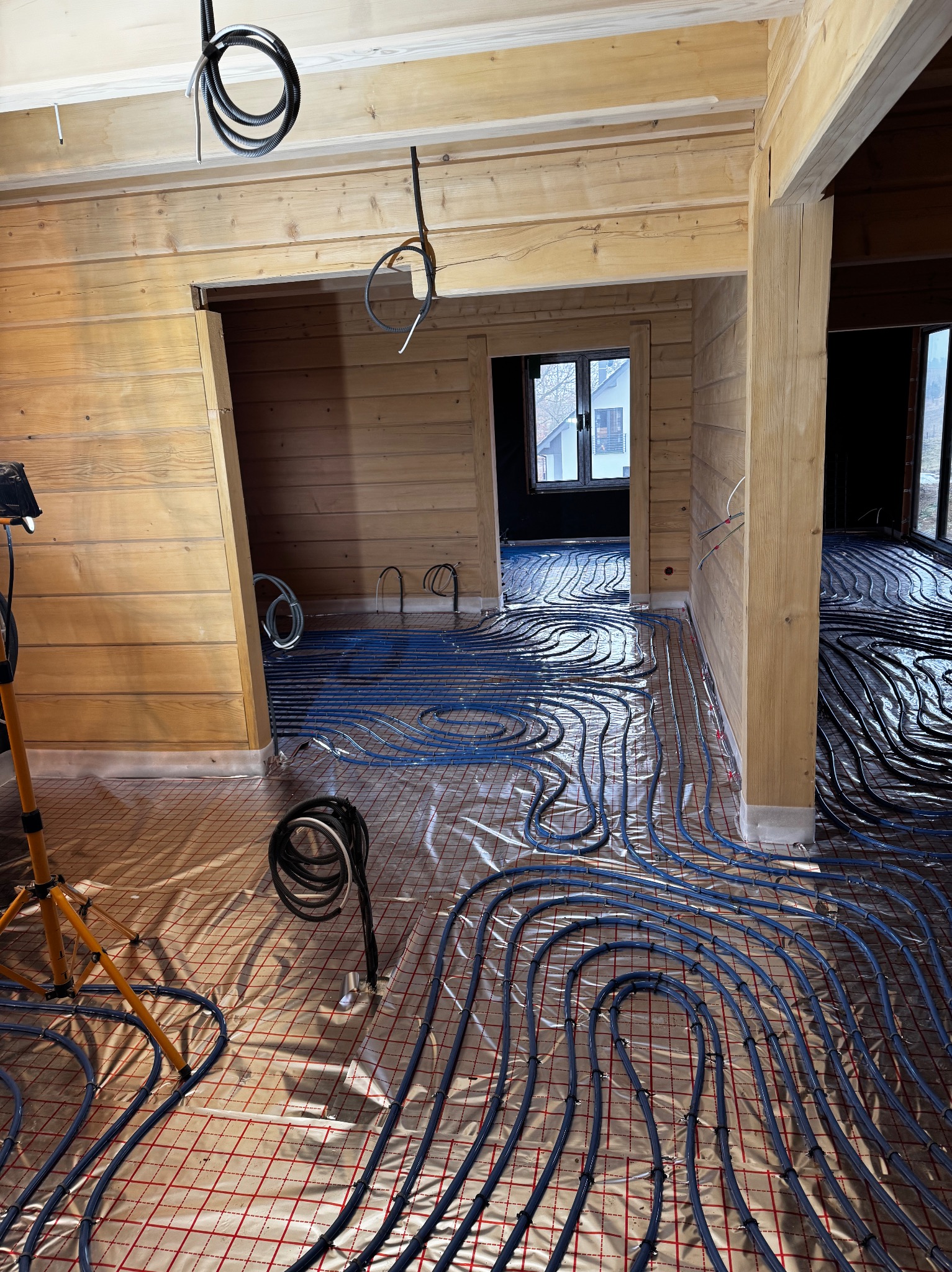 Wnętrze domu w trakcie budowy z widocznym systemem ogrzewania podłogowego w postaci niebieskich rur ułożonych na folii z czerwoną siatką, drewniane ściany i sufit w surowym stanie, widoczny przewód...