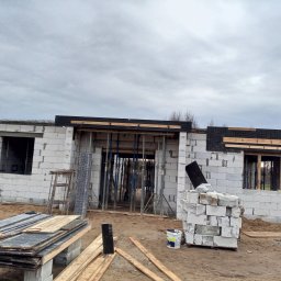 BM PROFIT - Wznoszenie ścian parterowego domu z betonu komórkowego, widoczne stalowe podpory szalunkowe i paleta z bloczkami, pochmurne niebo w tle.