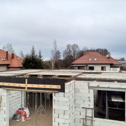 BM PROFIT - Wysoki kąt widzenia na budowę domu z bloczków, widoczna zbrojona płyta stropowa wsparta na stemplach, w tle domy jednorodzinne z czerwonymi dachami.