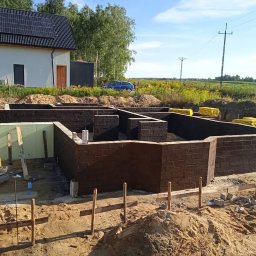 BM PROFIT - Wykopane fundamenty z wymurowanymi ścianami, zabezpieczone izolacją przeciwwilgociową, widoczne zielone płyty izolacyjne i żółte rolki papy, w tle dom z panelami słonecznymi.