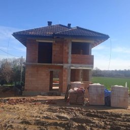 BM PROFIT - Surowy stan dwukondygnacyjnego domu z czerwonej cegły z dachem pokrytym dachówką, widok na plac budowy z błotnistym terenem, w tle zielone pole i turbiny wiatrowe.