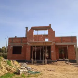 Budowa domu murowanego w trakcie wznoszenia ścian z czerwonej cegły, widoczne szalunki i rusztowania, teren budowy z hałdą piasku i gruzem.