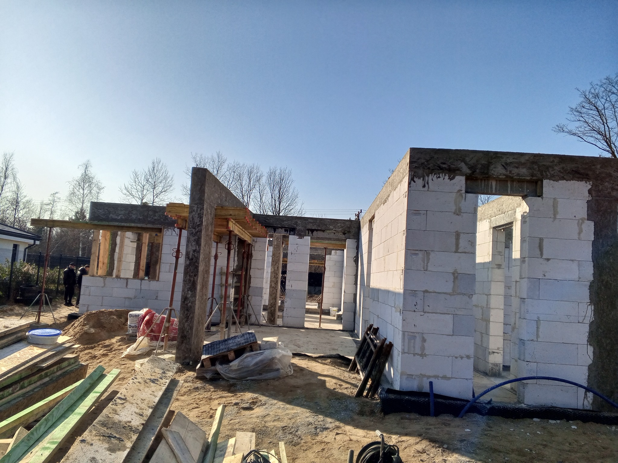 Wznoszenie ścian parterowego budynku z betonu komórkowego, widoczne drewniane szalunki okienne i stalowe podpory, na pierwszym planie deski i narzędzia budowlane, w tle pracujący robotnik.