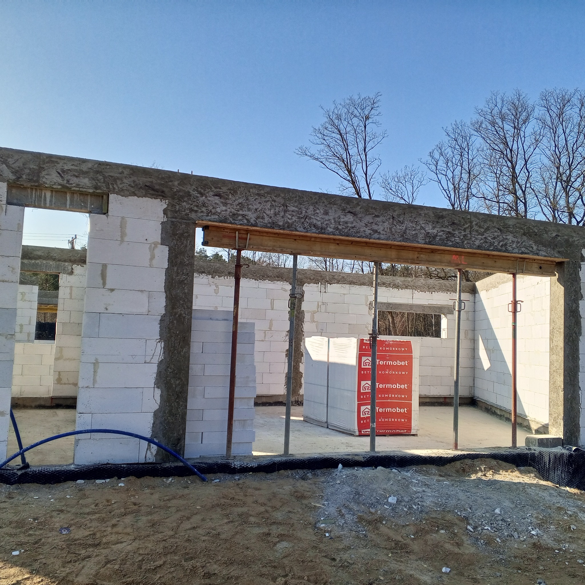 Budowa domu jednorodzinnego: ściany wzniesione z białych bloczków, widoczne stemple podtrzymujące nadproża, na zewnątrz paleta z materiałem budowlanym Termobet.