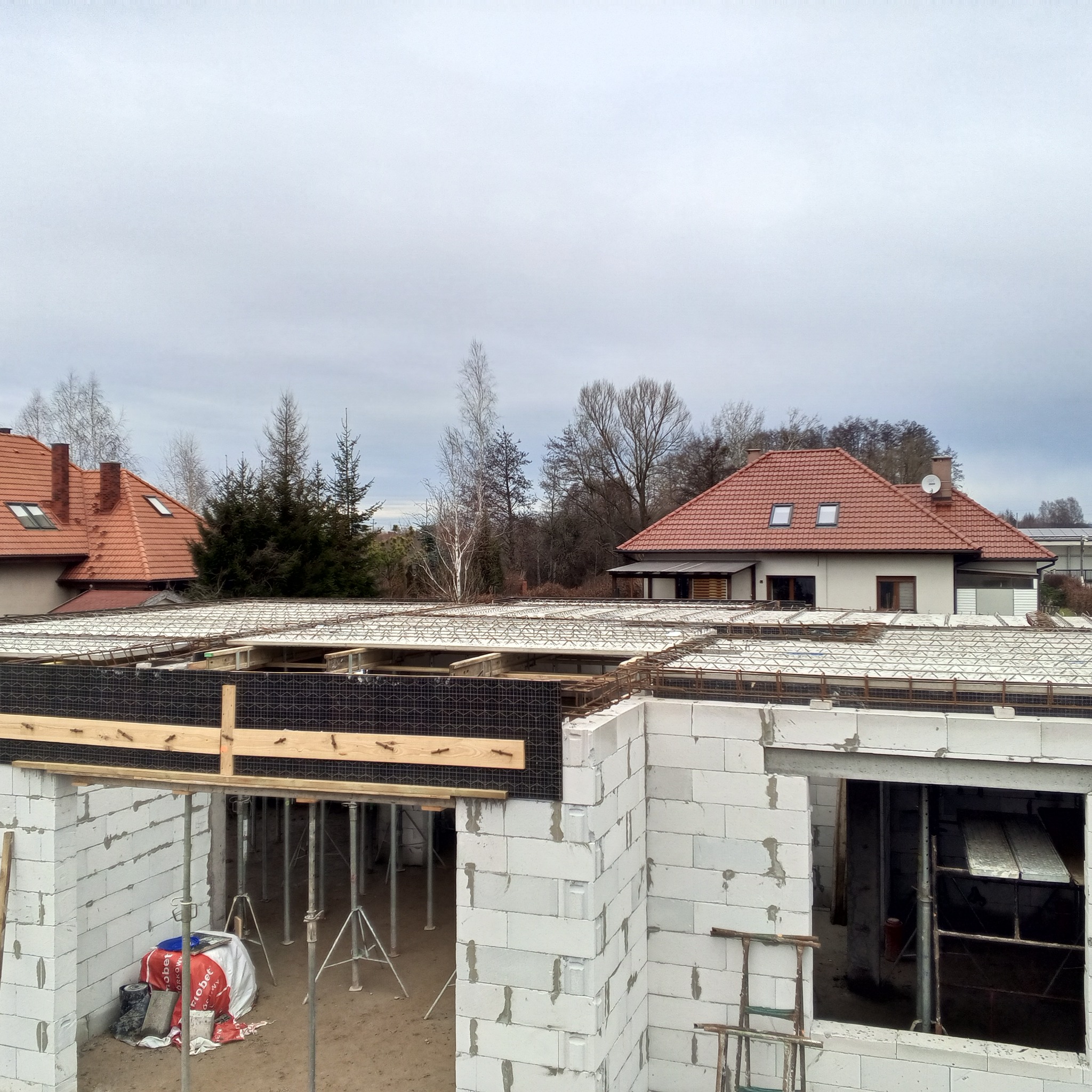 Wysoki kąt widzenia na budowę domu z bloczków, widoczna zbrojona płyta stropowa wsparta na stemplach, w tle domy jednorodzinne z czerwonymi dachami.