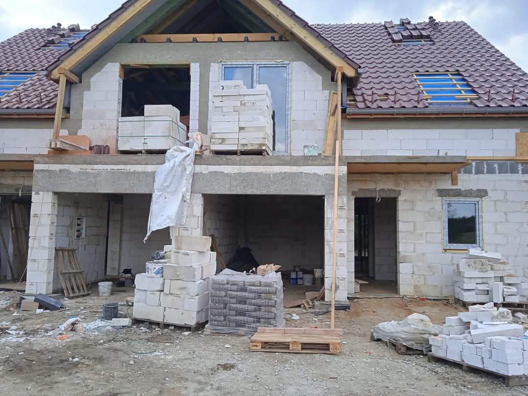 Surowy stan dwukondygnacyjnego budynku z białych bloczków, widoczne okna, częściowo położony dach z brązowej dachówki, na placu budowy porozrzucane materiały i narzędzia.