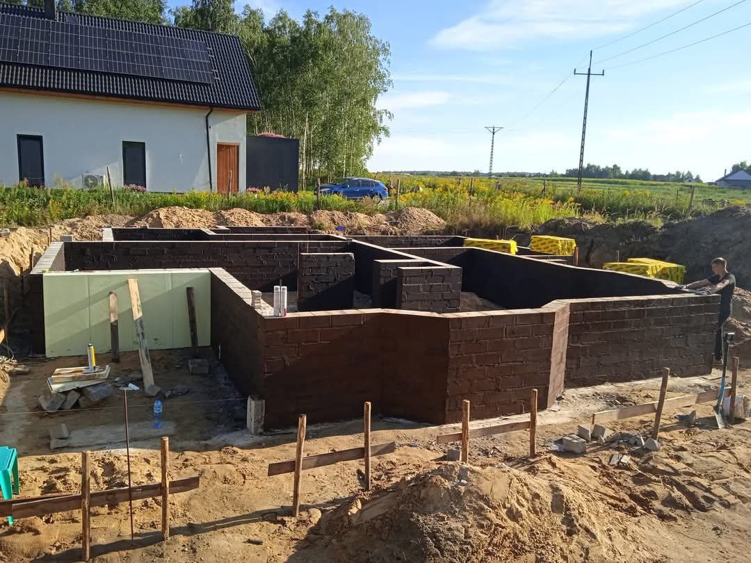Wykopane fundamenty z wymurowanymi ścianami, zabezpieczone izolacją przeciwwilgociową, widoczne zielone płyty izolacyjne i żółte rolki papy, w tle dom z panelami słonecznymi.