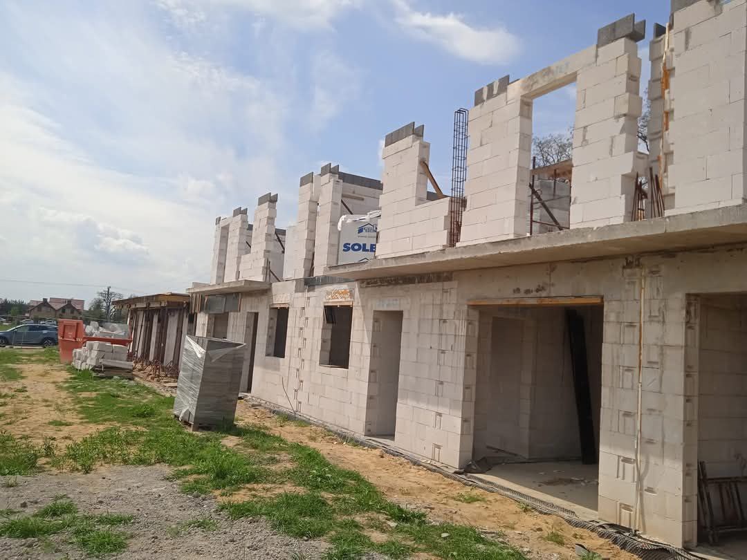 Wznoszenie ścian z betonu komórkowego na budowie szeregowca, widoczne zbrojenia i podciągi, palety z materiałem budowlanym obok.