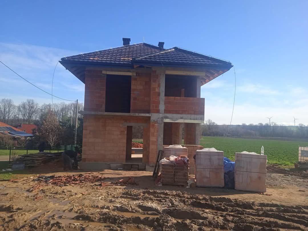 Surowy stan dwukondygnacyjnego domu z czerwonej cegły z dachem pokrytym dachówką, widok na plac budowy z błotnistym terenem, w tle zielone pole i turbiny wiatrowe.