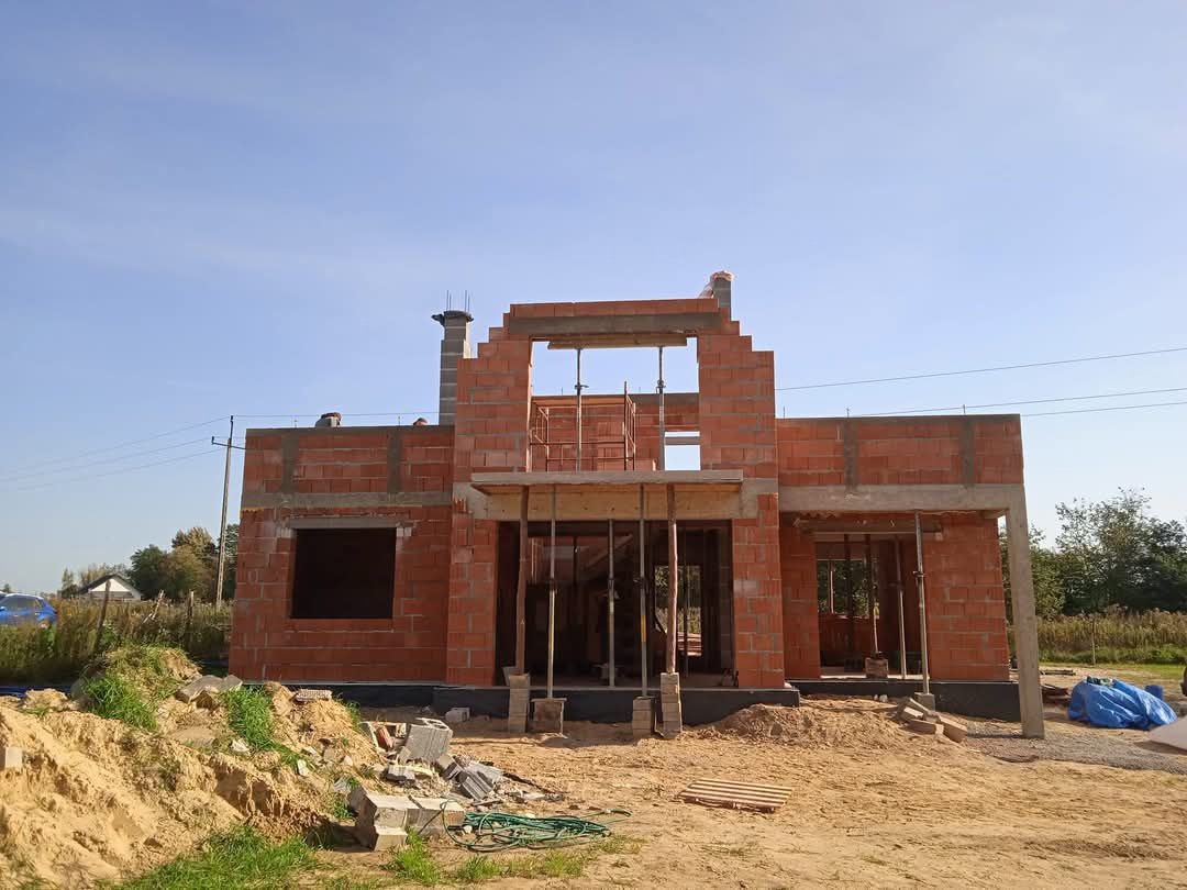 Budowa domu murowanego w trakcie wznoszenia ścian z czerwonej cegły, widoczne szalunki i rusztowania, teren budowy z hałdą piasku i gruzem.