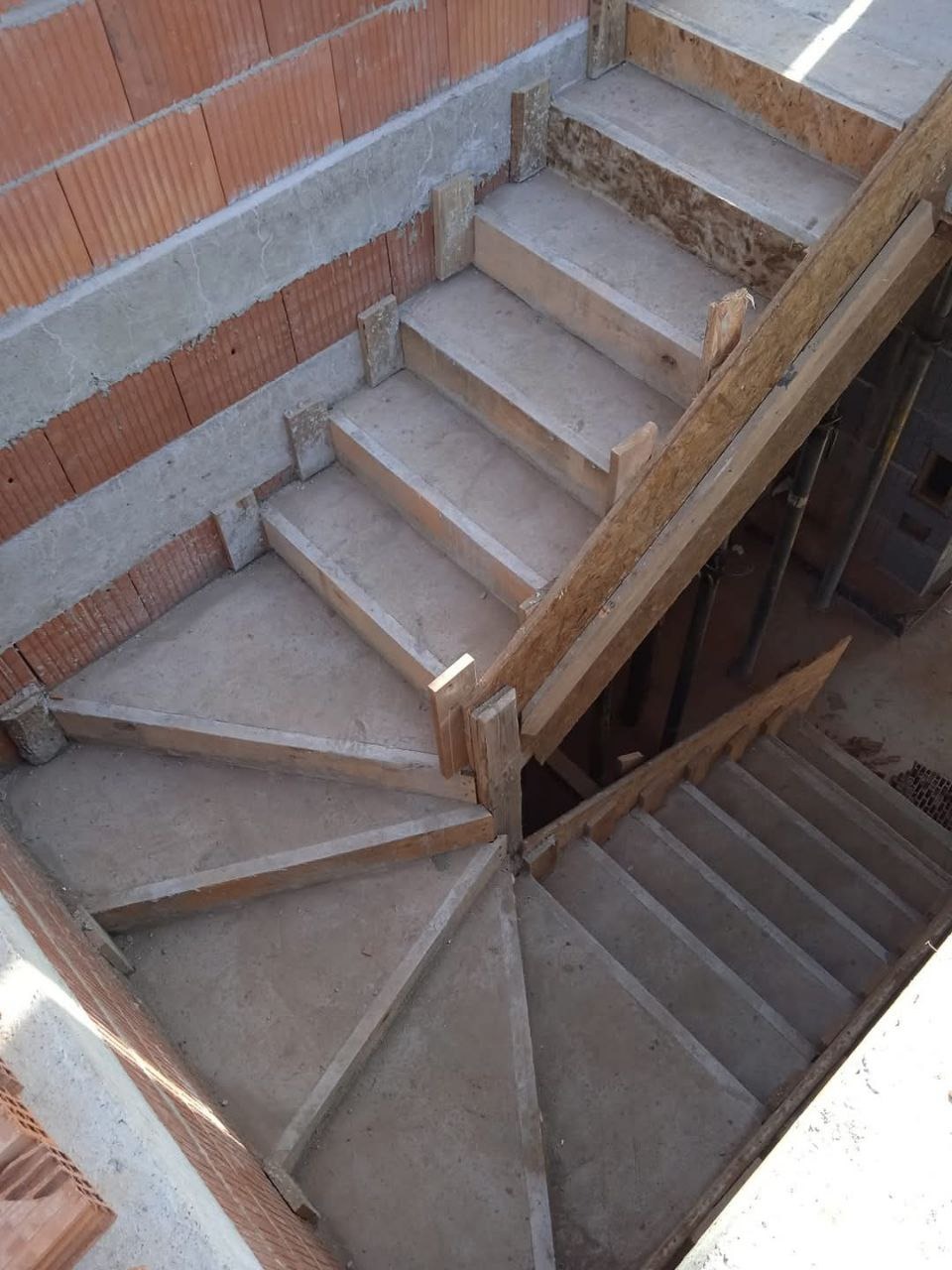 Surowe schody żelbetowe w trakcie budowy, z widocznym szalunkiem drewnianym i fragmentem murowanej ściany z pustaków.