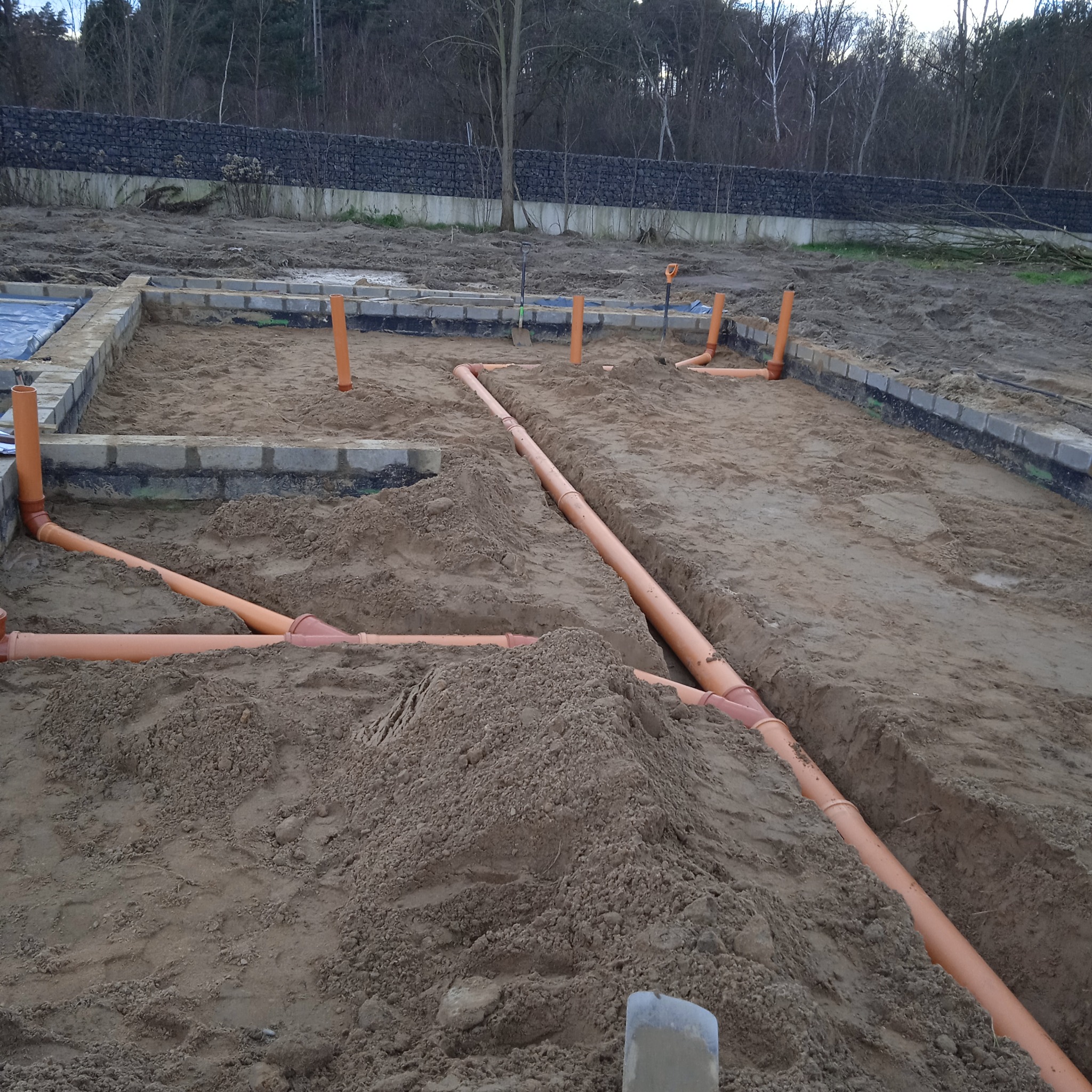 Przygotowanie fundamentów pod budynek: widoczne ułożone rury kanalizacyjne w wykopie, obrys fundamentów z bloczków betonowych, teren budowy z ziemią i drzewami w tle.