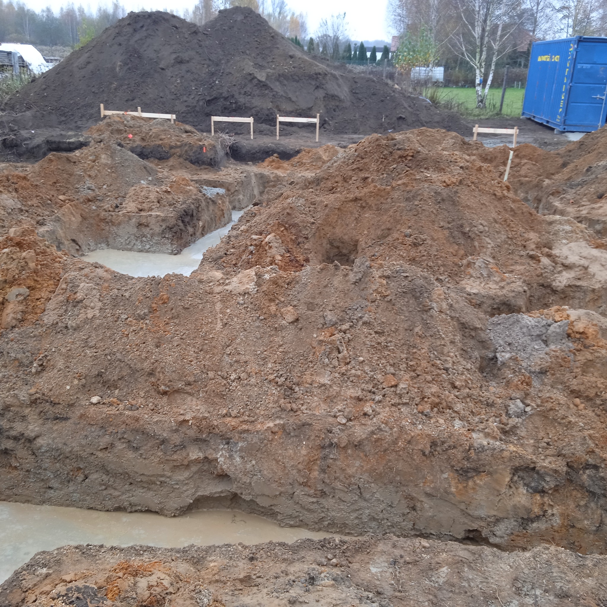 Wykopy pod fundamenty zalane wodą, widoczne drewniane szalunki i hałda ziemi w tle, obok kontener budowlany.
