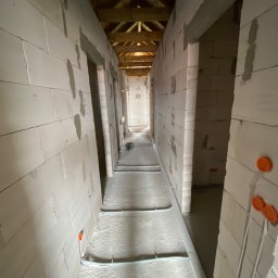 ElMac Maciej Lewandowski - Wnętrze w trakcie wykończenia: surowe ściany z betonu komórkowego, instalacja ogrzewania podłogowego z rurkami i rozdzielaczem, drewniana konstrukcja dachu.