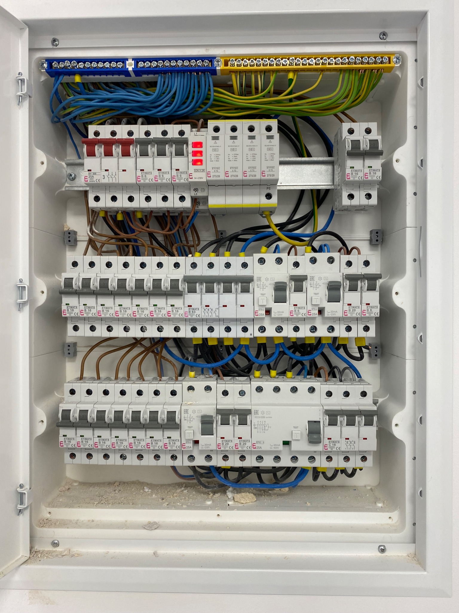 Rozdzielnica elektryczna w białej obudowie, widoczne bezpieczniki, kable w różnych kolorach (niebieski, brązowy, zielono-żółty). Instalacja po montażu, gotowa do użytku.