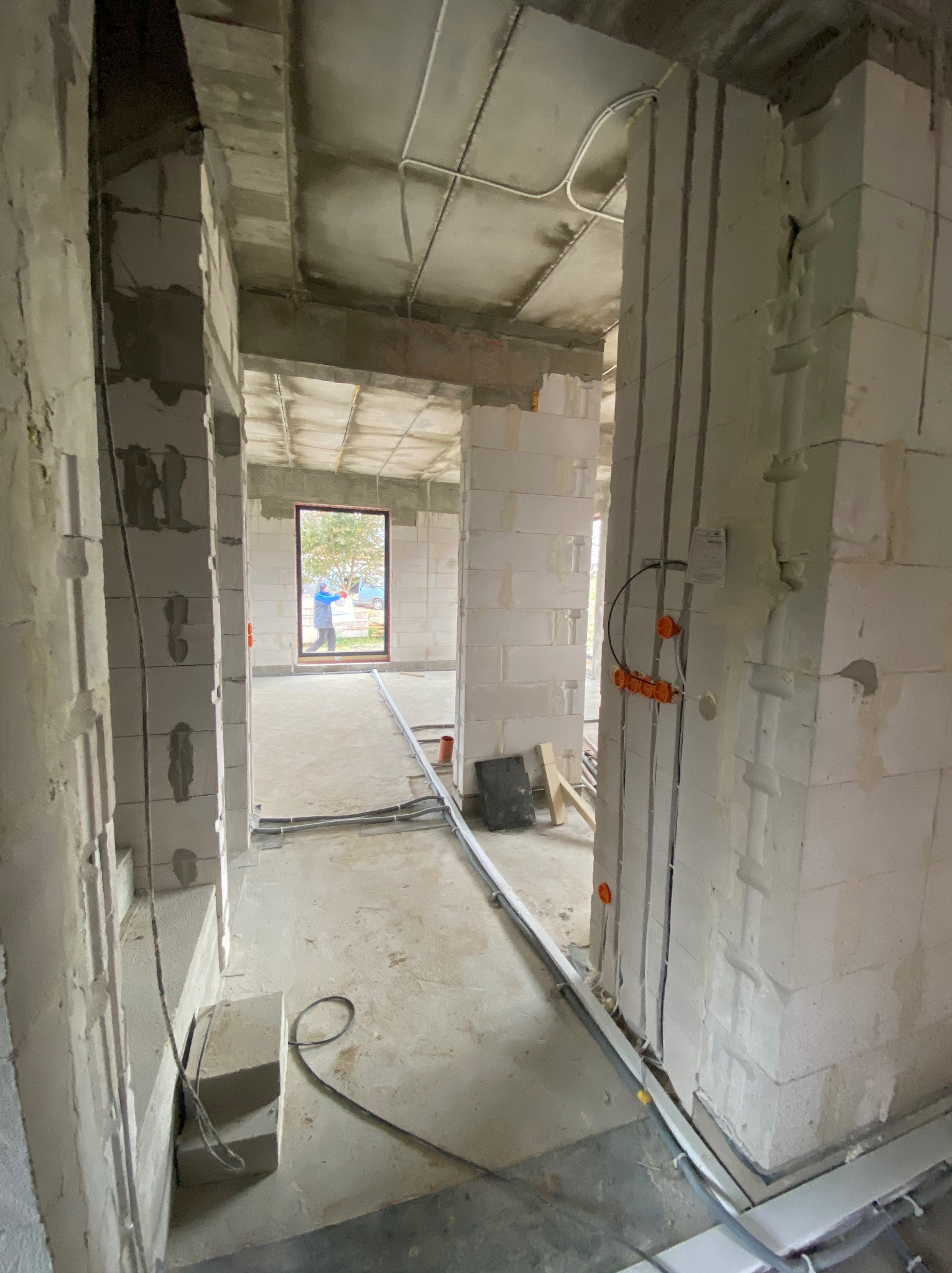 Wnętrze w stanie surowym z widoczną instalacją elektryczną w ścianach i na podłodze. Widoczne puszki elektryczne i przewody. W tle okno z widokiem na zewnątrz.