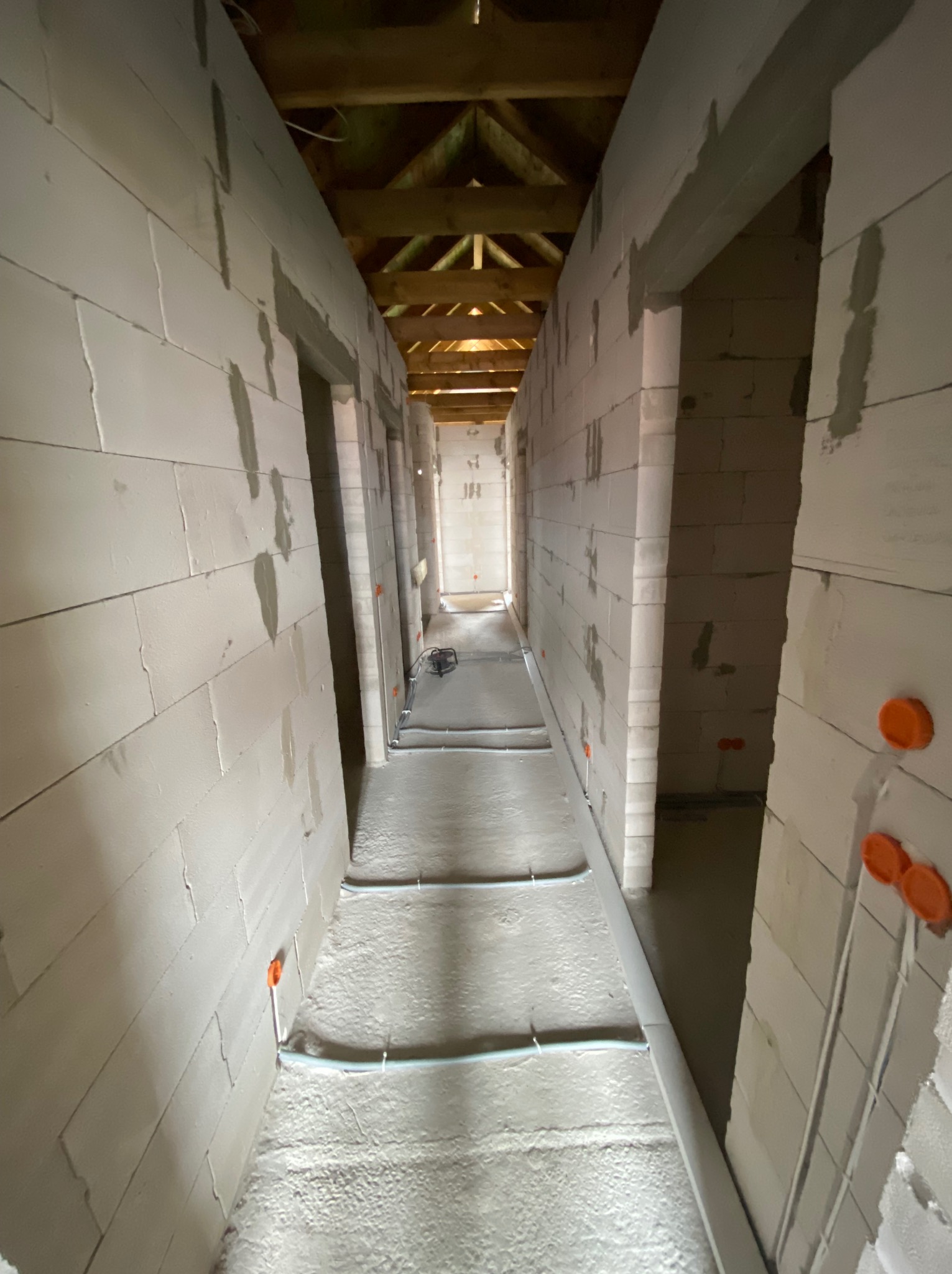 Wnętrze w trakcie wykończenia: surowe ściany z betonu komórkowego, instalacja ogrzewania podłogowego z rurkami i rozdzielaczem, drewniana konstrukcja dachu.