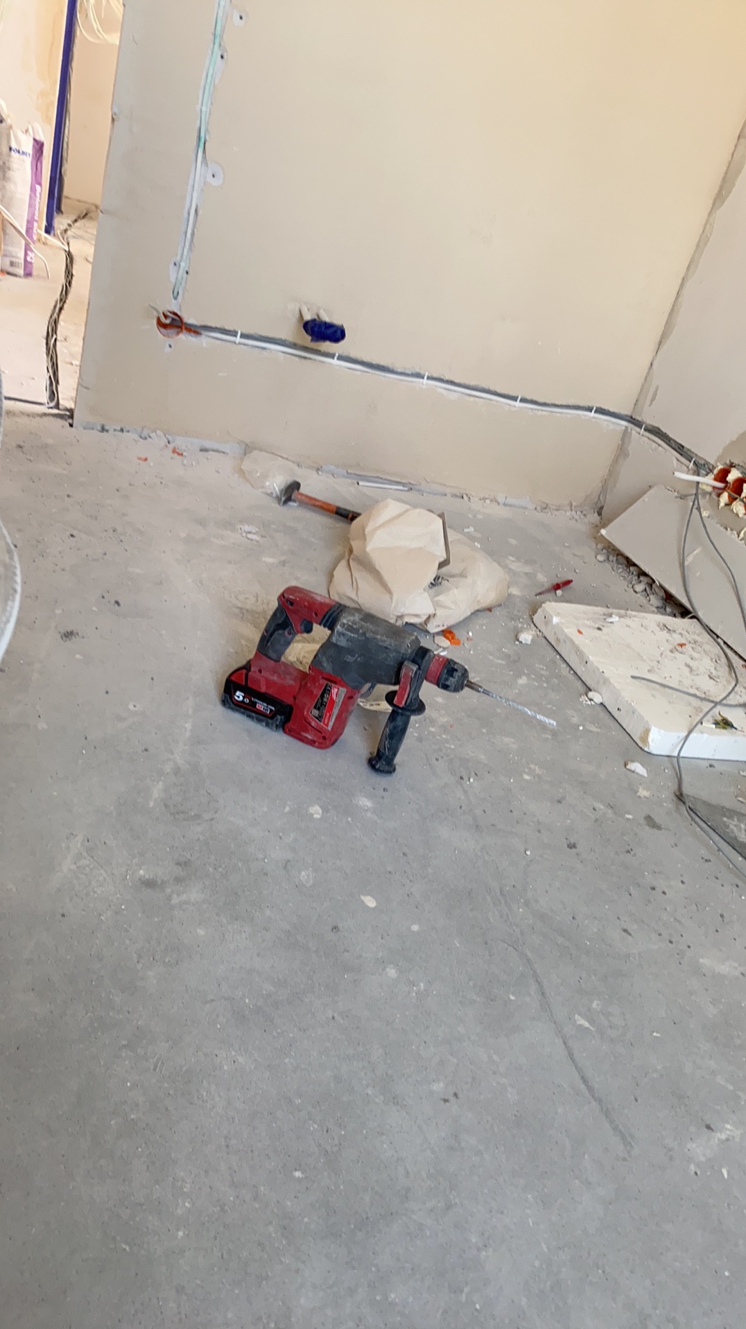 Czerwony młotowiertarka akumulatorowa na szarym podłożu, w tle ściana z widocznymi przewodami elektrycznymi i gładzią. Etap prac wykończeniowych.