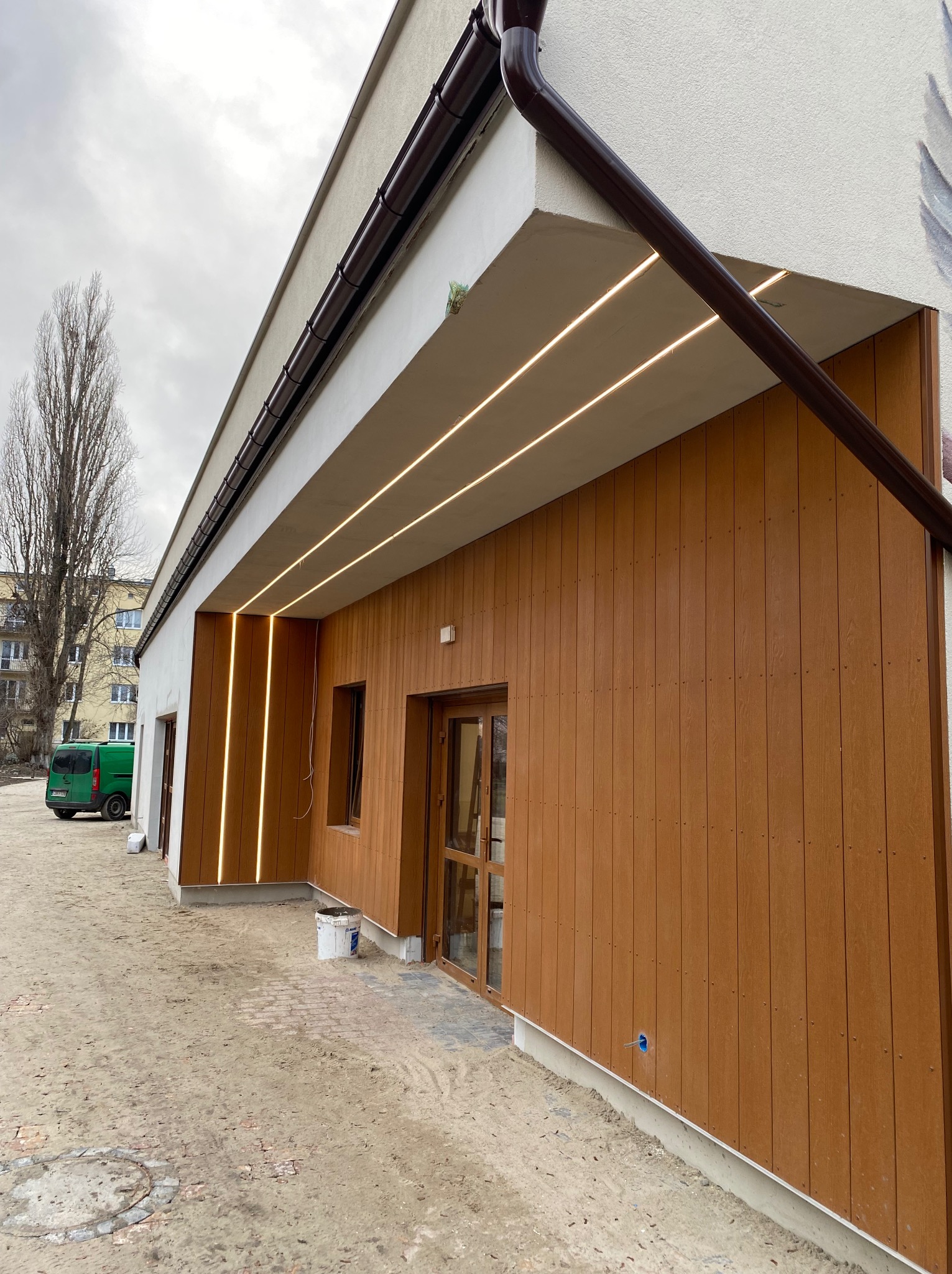 Elewacja budynku z drewnianą okładziną i liniowym oświetleniem LED pod okapem. Widoczny fragment rynny i teren wokół budynku w trakcie prac wykończeniowych.