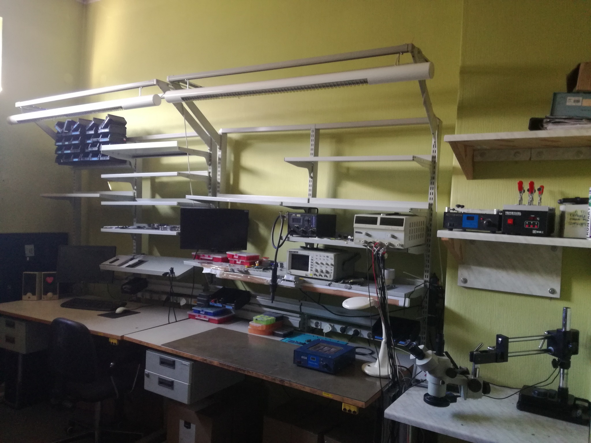 Stanowisko serwisowe z oscyloskopem, mikroskopem i innymi narzędziami diagnostycznymi na blacie roboczym, oświetlone lampą jarzeniową i z półkami pełnymi komponentów.
