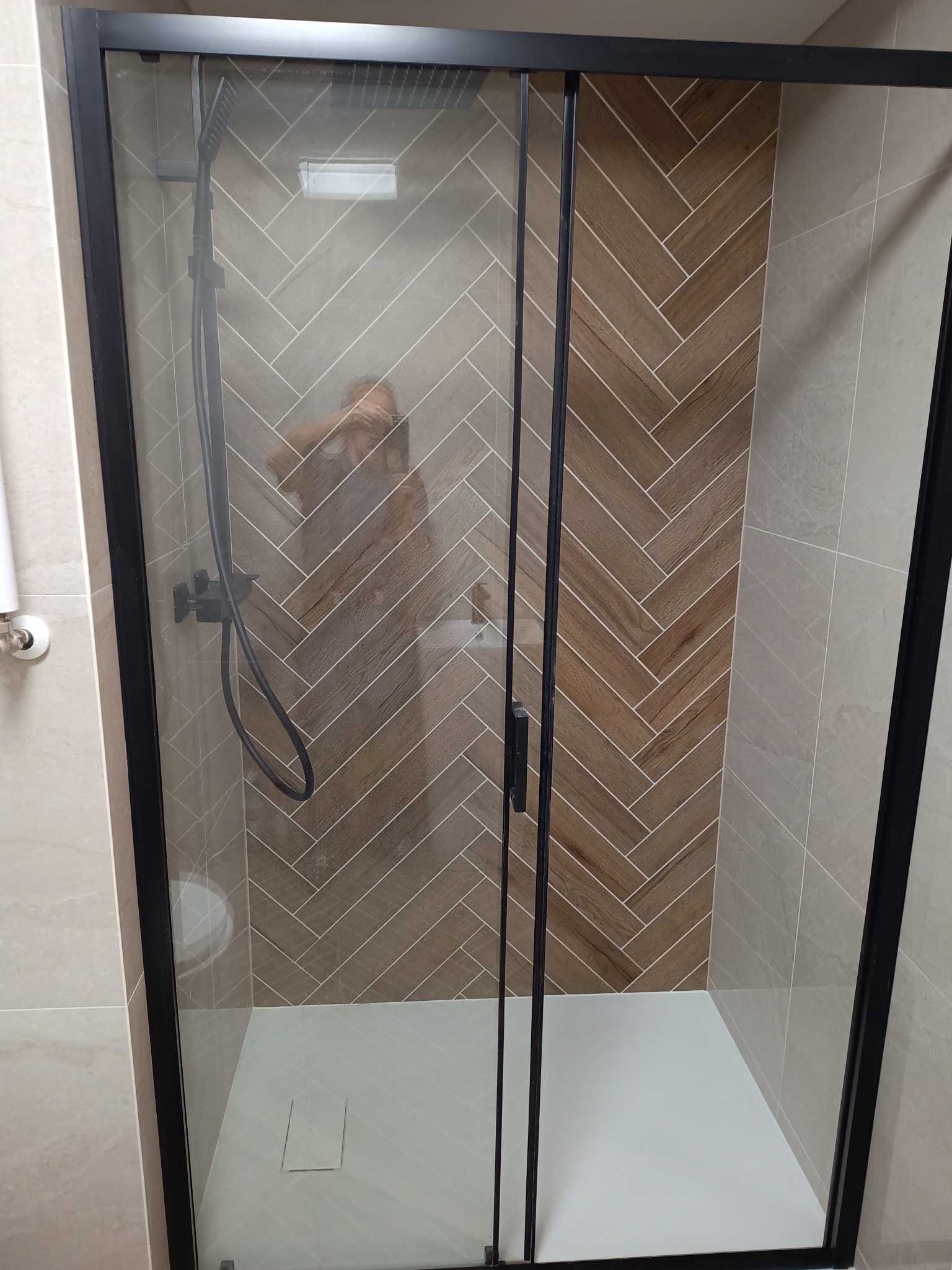 Kabina prysznicowa z czarnymi ramami i drzwiami przesuwnymi, wykończona płytkami w jodełkę w odcieniach brązu i beżu, z widoczną białą brodzikiem i czarną armaturą.