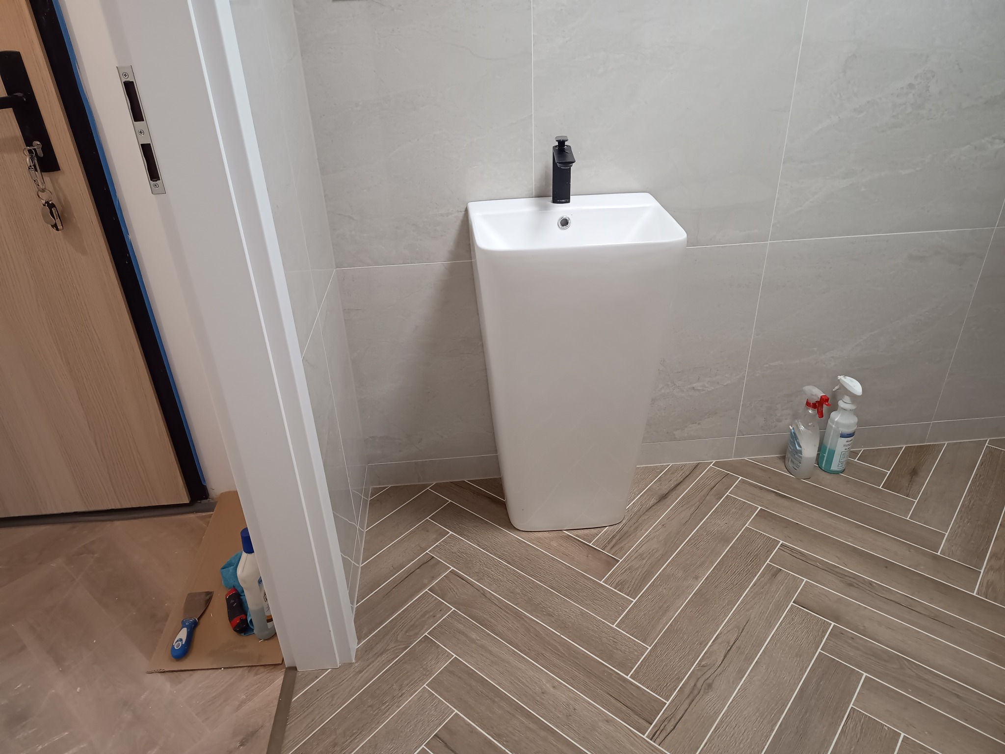 Biała, wolnostojąca umywalka z czarną armaturą na tle szarych płytek ściennych i podłogi ułożonej w jodełkę; widoczne środki czystości i narzędzia przy drzwiach.