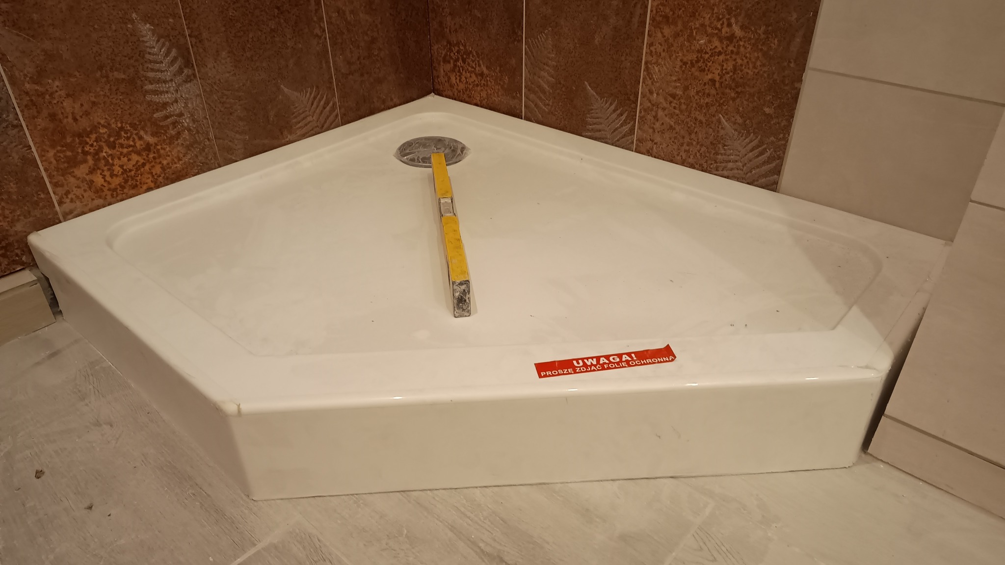 Biały brodzik narożny z naklejką ostrzegawczą 'Uwaga! Proszę zdjąć folię ochronną', poziomica leżąca na dnie, tło z brązowych płytek z motywem liści paproci.