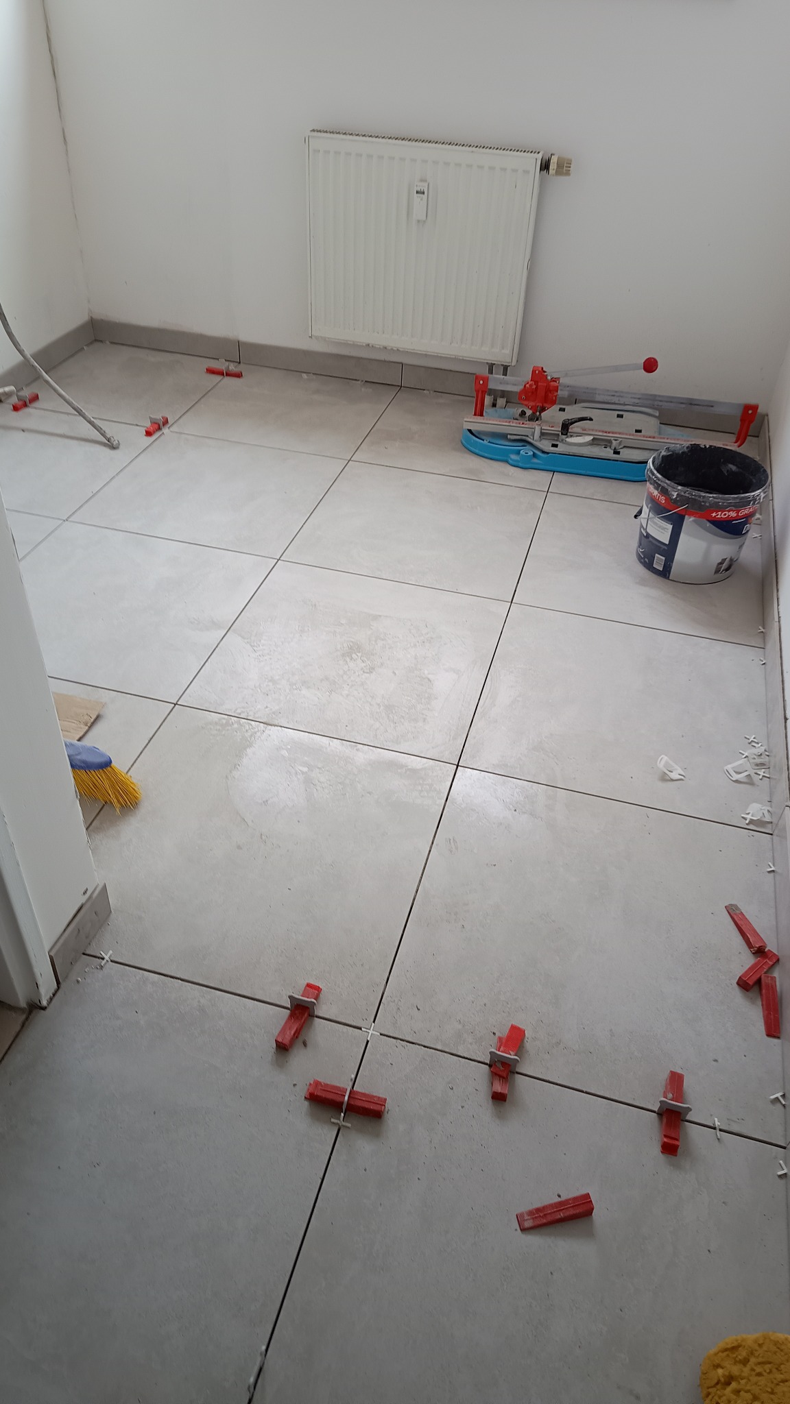 Układanie jasnoszarych płytek podłogowych z użyciem czerwonych klipsów poziomujących; w tle widoczna przecinarka do płytek, wiadro z klejem i grzejnik.