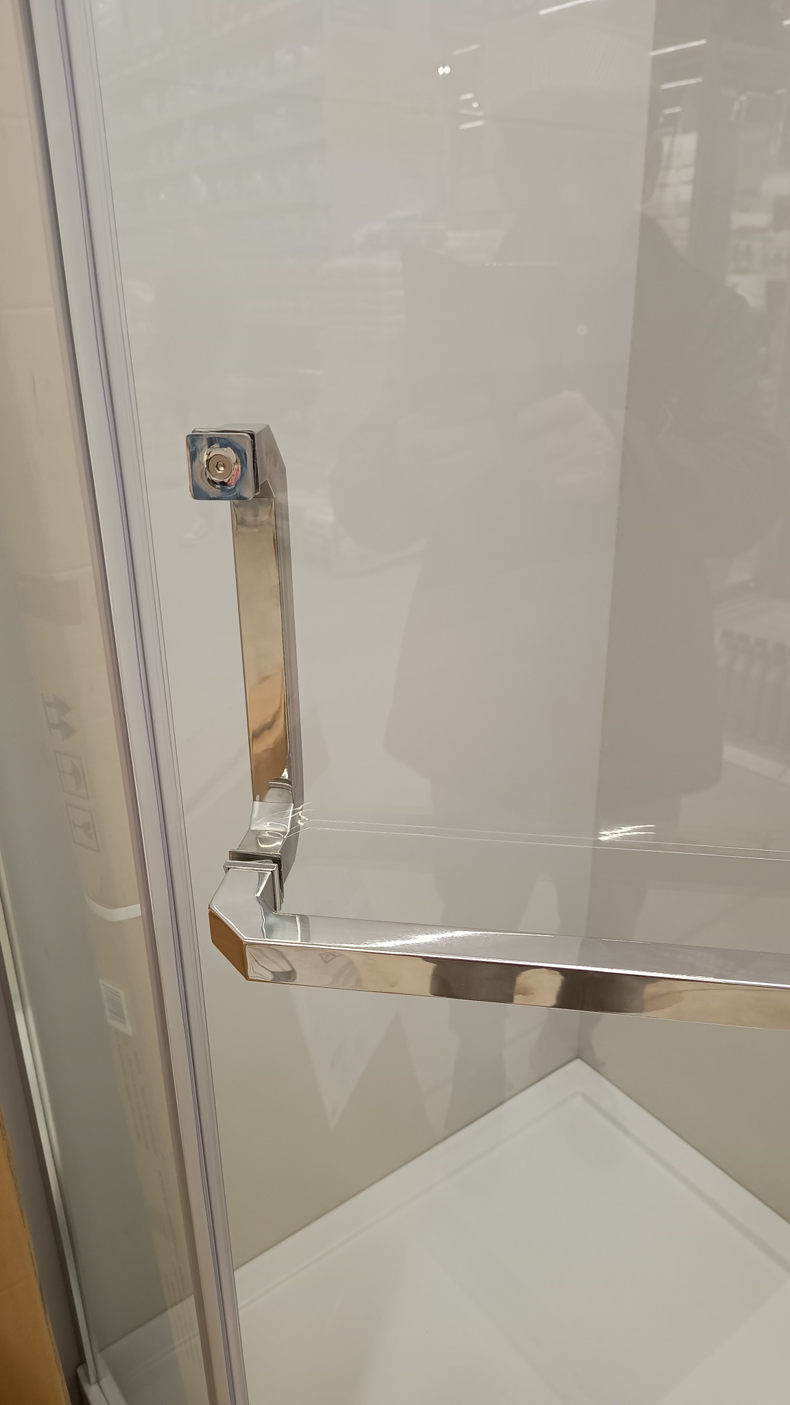 Szczegół chromowanego uchwytu kabiny prysznicowej z widocznym zamkiem, fragment białego brodzika w tle.