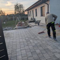 FAVOR - ANDRII SKLIAR - Pracownik zamiata piasek na nowo ułożonej, szarej kostce brukowej na podjeździe. Widoczny fragment ogrodzenia, domu i narzędzia pracy.