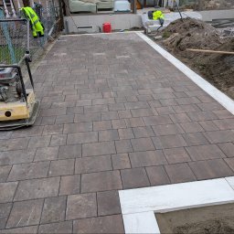 FAVOR - ANDRII SKLIAR - Ułożona, brązowa kostka brukowa z białym obramowaniem. Na pierwszym planie ubijarka. W tle widoczny teren przygotowany pod dalsze prace.