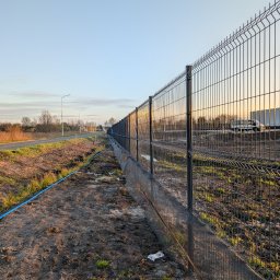 FAVOR - ANDRII SKLIAR - Długi, panelowy, metalowy płot z siatki, zamontowany wzdłuż drogi. Widoczny fragment drogi i pobocze z niebieskim wężem. W tle budynki i pojazd.